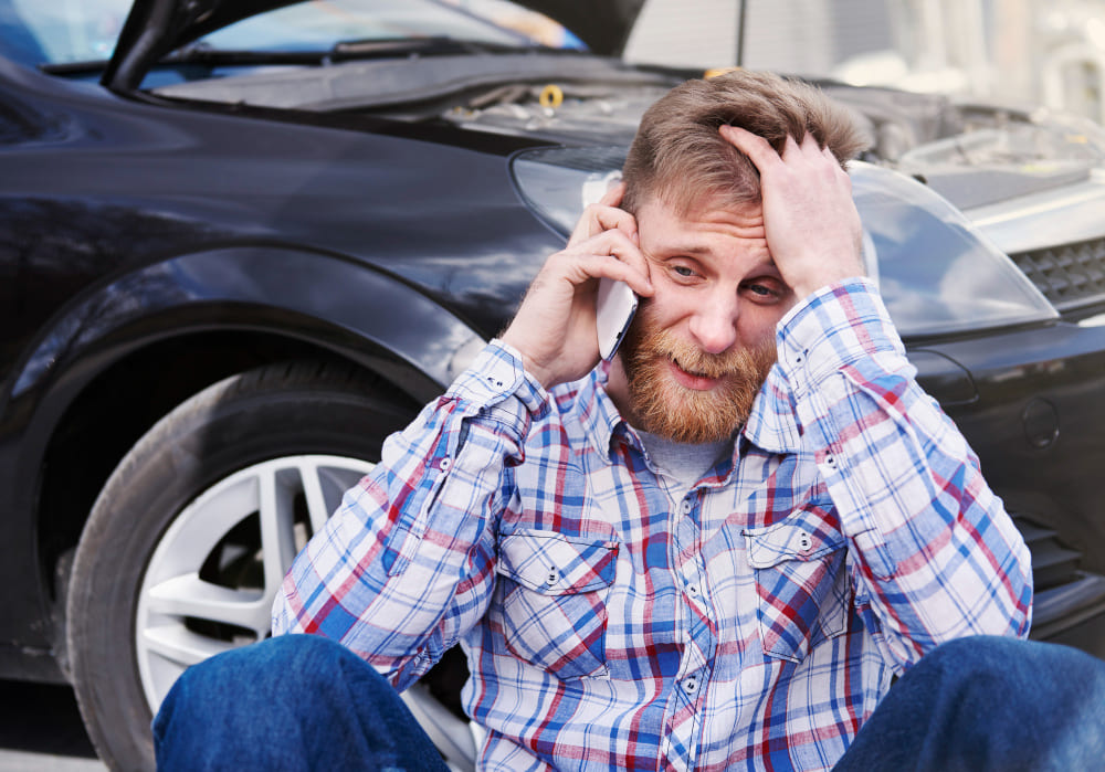 stressed-driver-calling-for-help-after-car-breakdown Frustrated man sitting on the ground beside a broken-down car