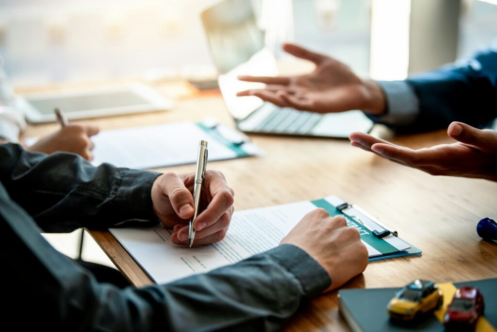 Person signing a car insurance contract on a clipboard during a meeting with an agent, toy cars visible on the desk