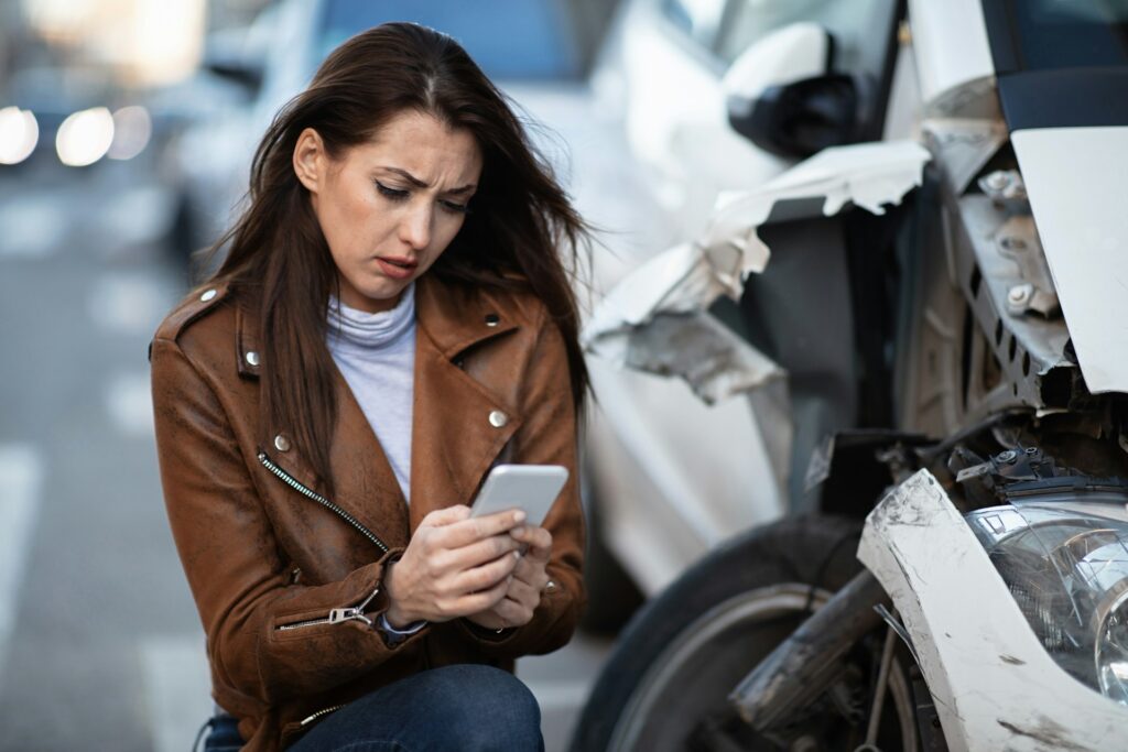 Distressed woman using smartphone to report a car accident while crouching next to a damaged vehicle